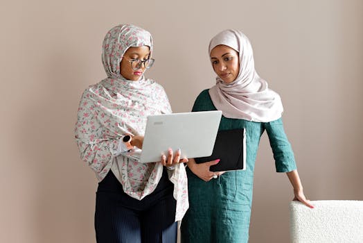Two women wearing headscarves working on a laptop in an office setting. Perfect for business diversity themes.