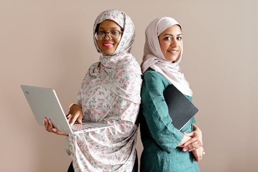 Two Muslim businesswomen in hijabs standing confidently with a laptop and notebook. Professional and modern representation.