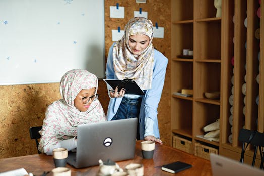 Two women in hijabs working together in a modern office setting with laptops and notes.
