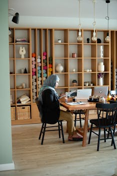 A professional Muslim woman in a hijab working on laptops in a stylish office with wooden shelves.