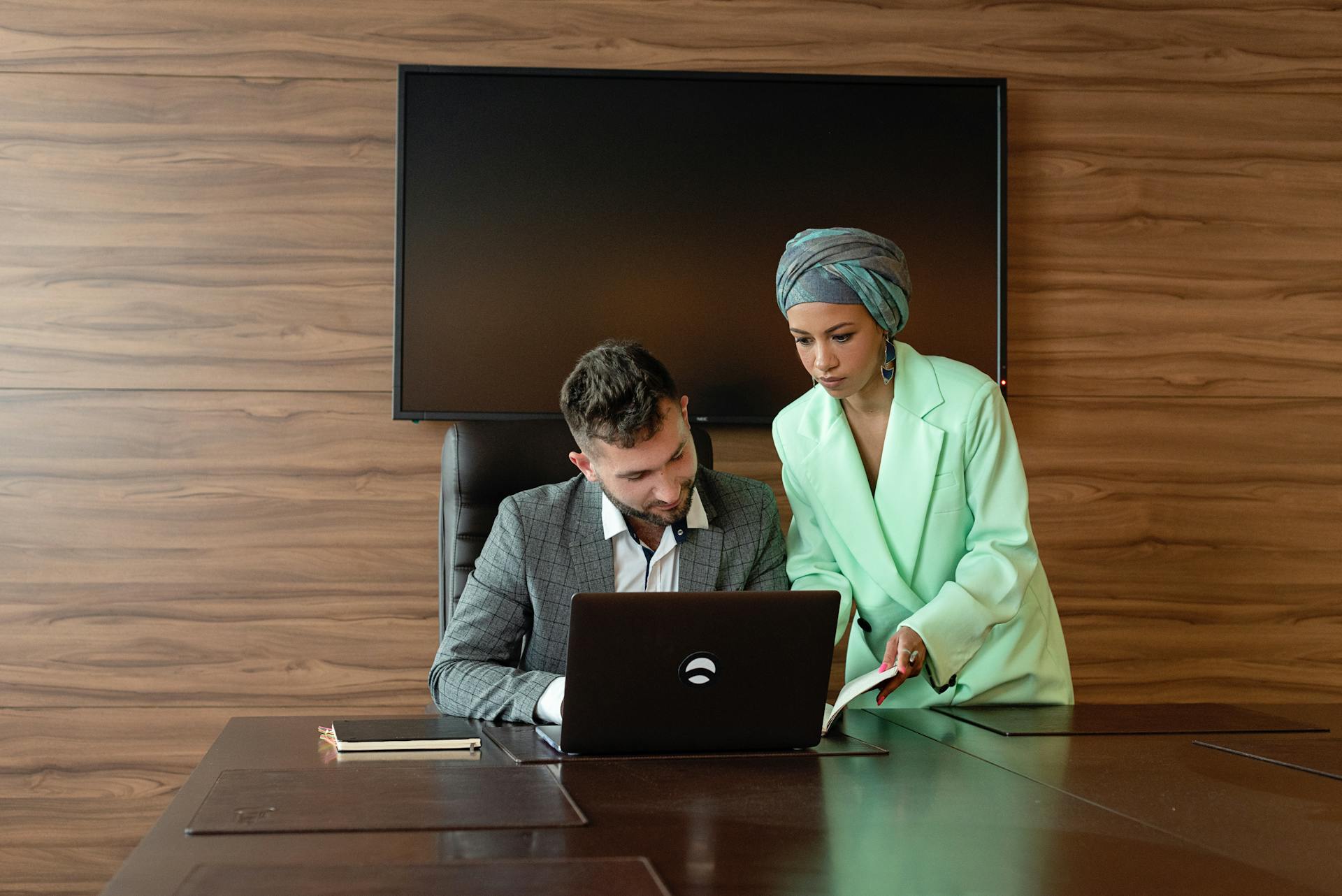 Two business professionals collaborate at a desk with a laptop in a modern office setting.