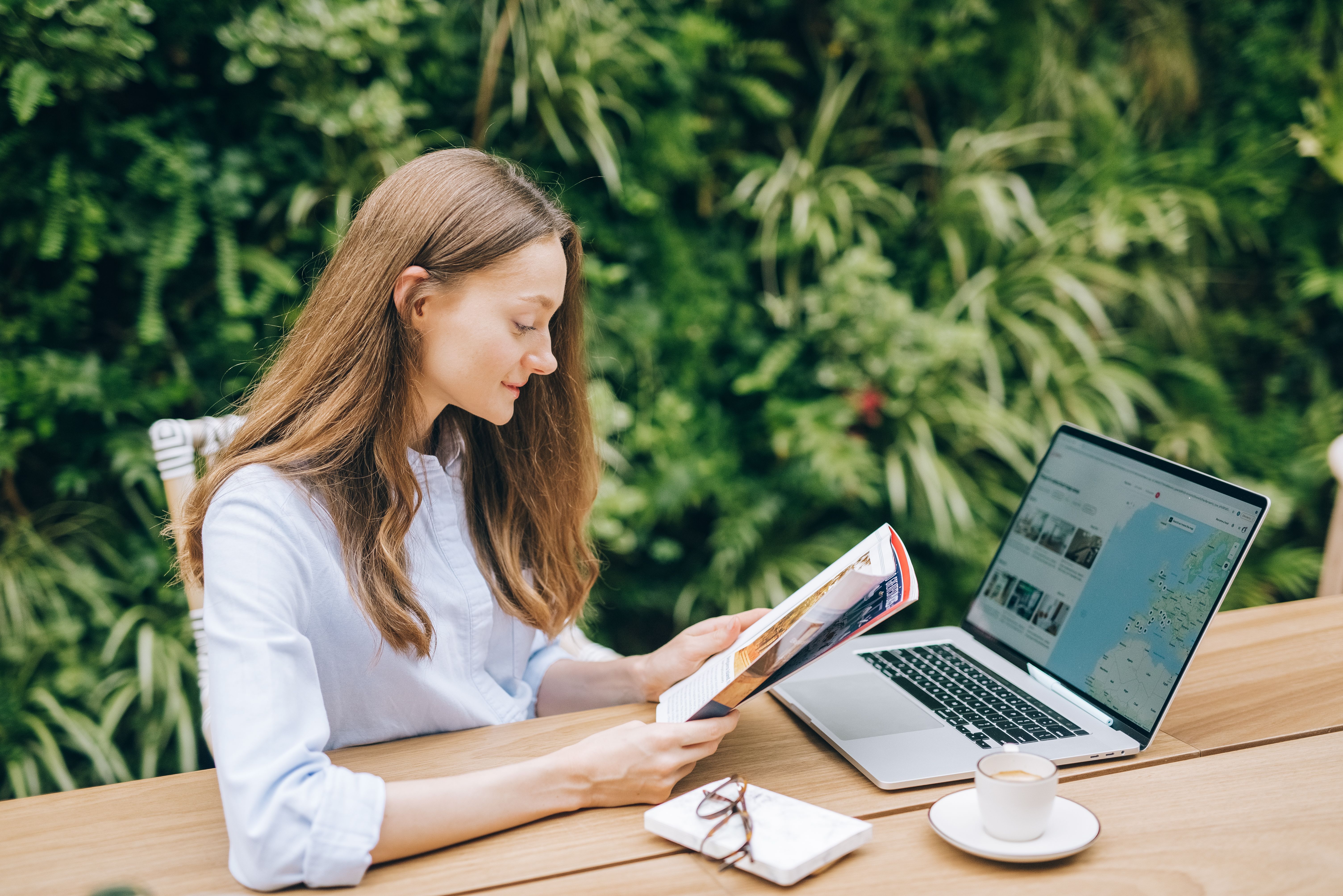 Caucasian woman reading a magazine at a wooden table with a laptop, outdoors.