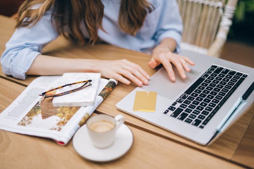 Woman working on a laptop with coffee and a magazine indoors, featuring a casual setting.