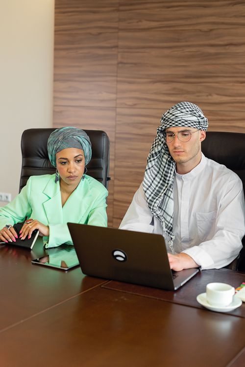 Free Man and woman in traditional attire collaborating over a laptop in a modern office setting. Stock Photo