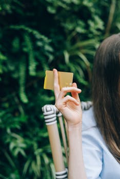 A woman's hand holding a yellow card, set against a lush garden backdrop.