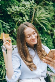 Happy woman using smartphone and holding credit card in lush garden setting, enjoying online shopping.