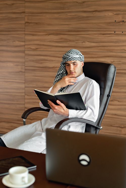 Free Businessman in traditional attire reads a document in a modern office setting. Stock Photo