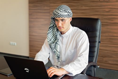 Free Middle Eastern man in traditional attire working on a laptop in an office setting. Stock Photo