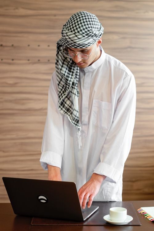 Free Middle Eastern man in traditional attire working on a laptop in an office setting. Stock Photo