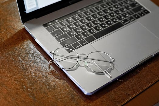 Close-up of round eyeglasses placed on a laptop keyboard, atop a brown leather surface.