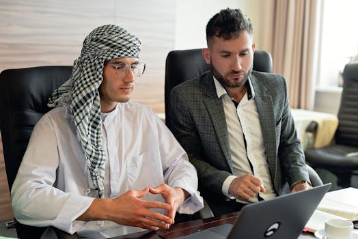Two businessmen in suits collaborate on a laptop in a modern office setting, focused and engaged.