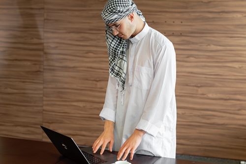 Free Middle Eastern man in traditional attire working on a laptop indoors. Stock Photo