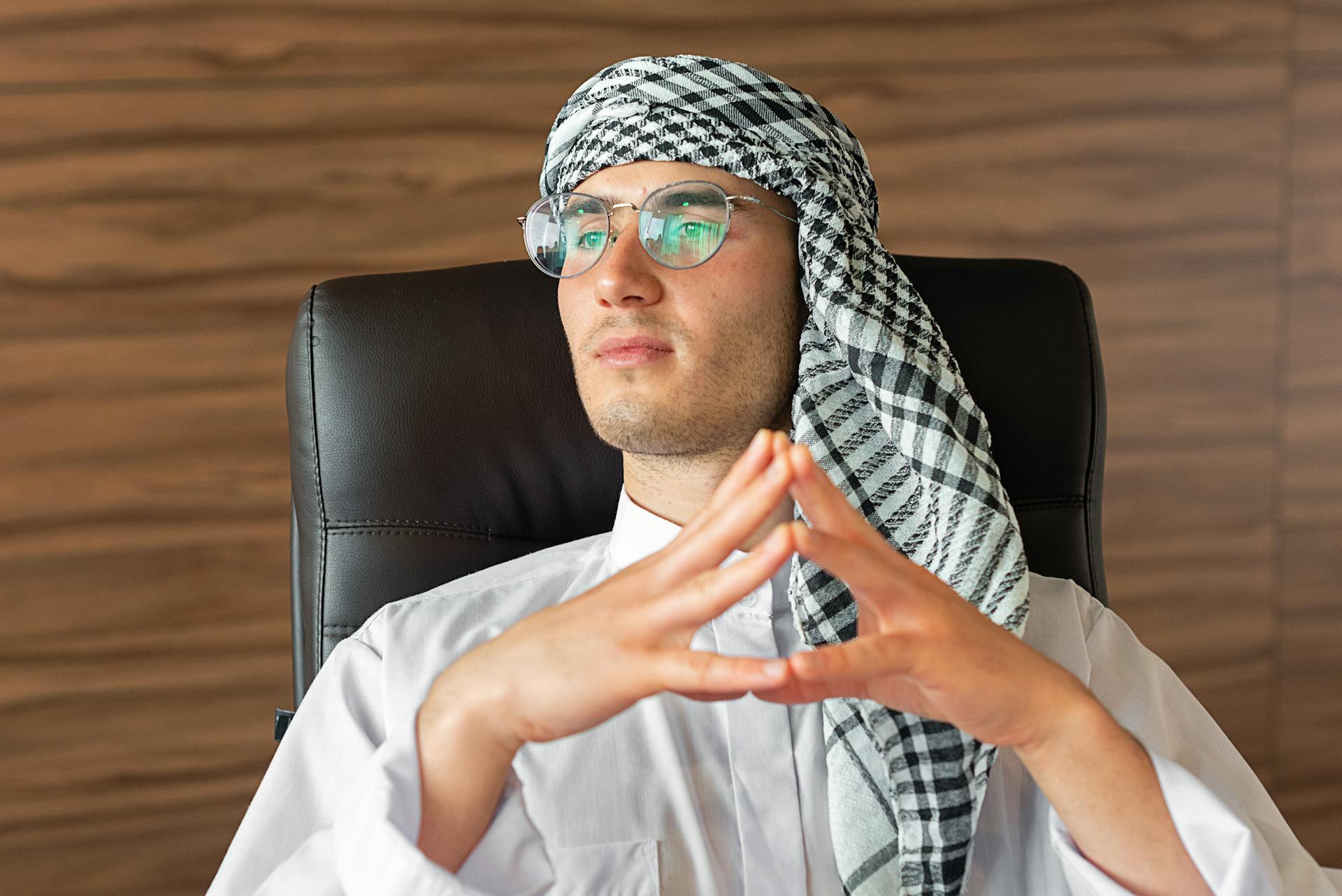 Middle Eastern man in professional attire with headscarf and glasses seated in a modern office environment.