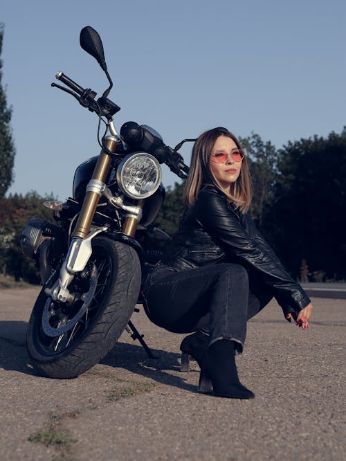 Woman in Black Leather Jacket and Black Pants Sitting Beside a Black Motorcycle
