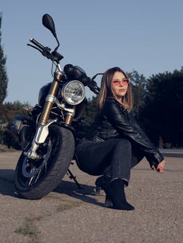 Confident woman in leather jacket poses with motorcycle outdoors under clear sky.