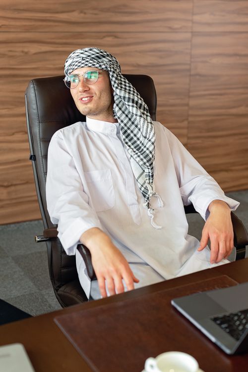 Free Middle Eastern man in traditional attire sitting relaxed in a modern office chair. Stock Photo