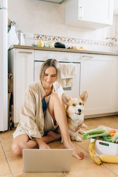 Young woman working on laptop in kitchen with friendly Corgi beside her, symbolizing remote work lifestyle.