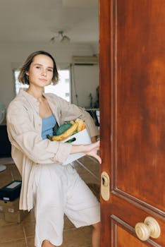 Young woman standing in doorway holding box of fresh groceries, smiling indoors.