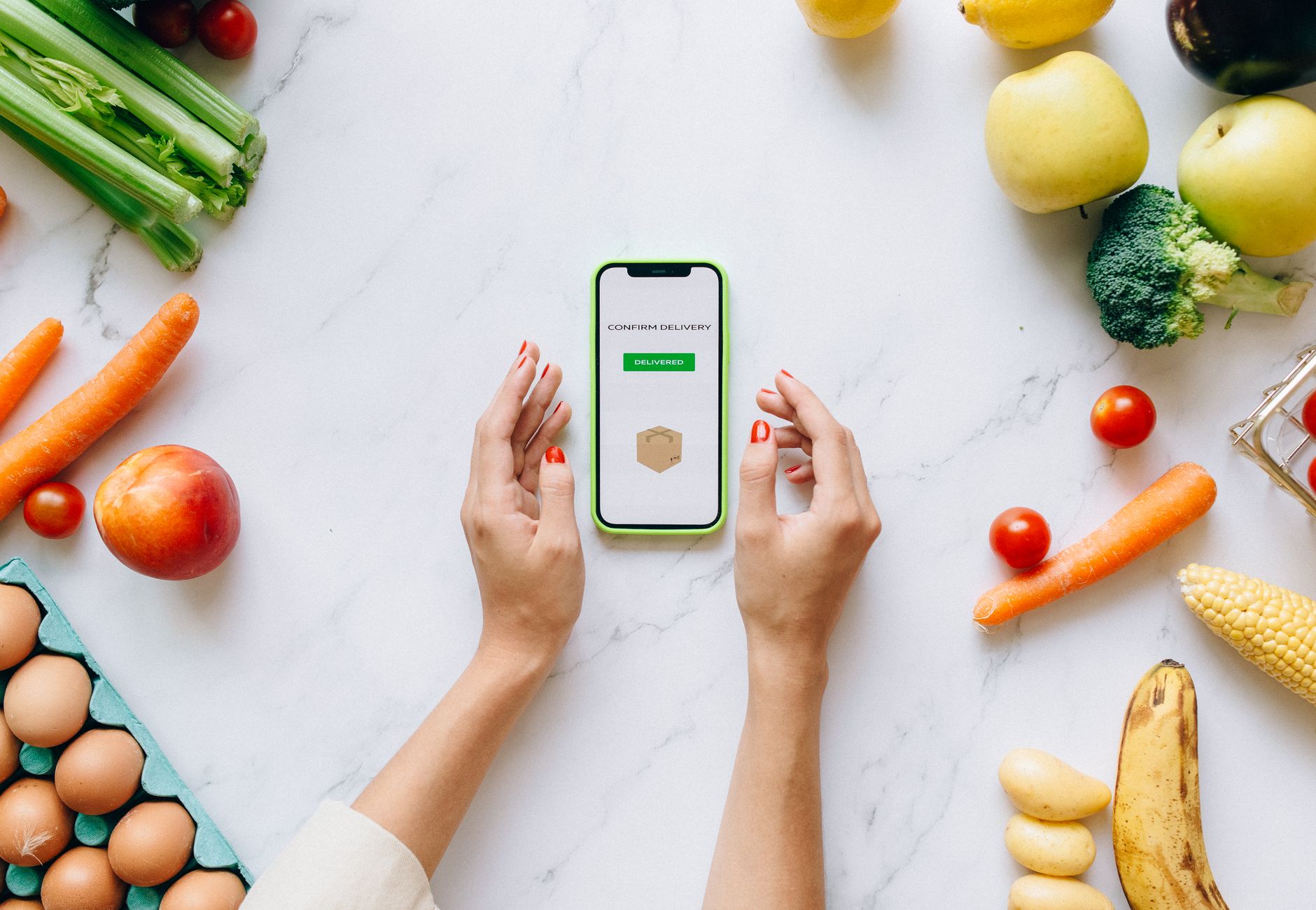 Hands holding smartphone displaying online grocery order amidst fresh vegetables and fruits.