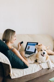Woman with laptop and corgi dog shopping online with credit card on sofa.