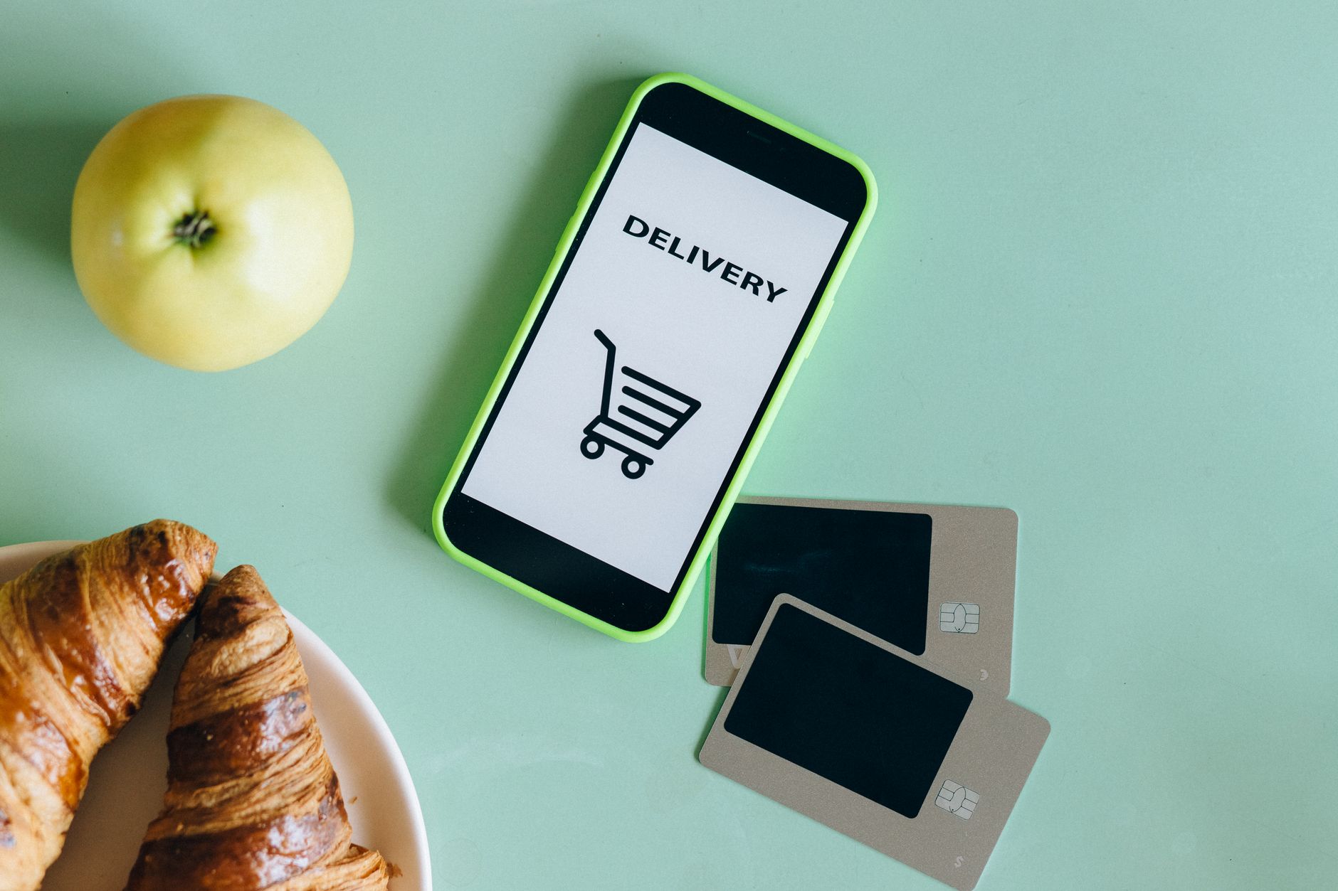 A smartphone with a delivery app next to an apple, croissants, and credit cards on a green surface.