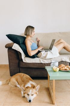 A woman sits on a sofa using a laptop, with her corgi dog resting nearby, in a cozy indoor setting.