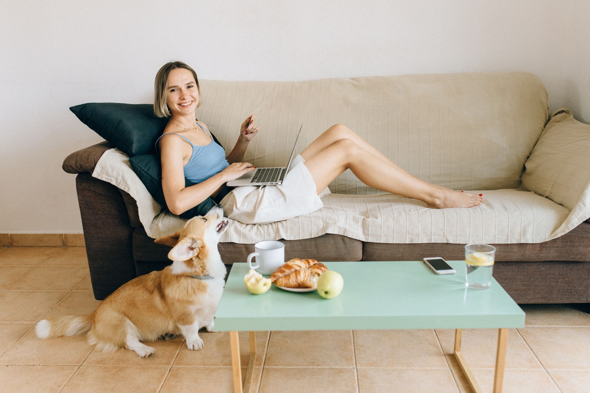 Woman relaxing on a sofa with a laptop, accompanied by a cute corgi dog. Cozy lifestyle indoors.