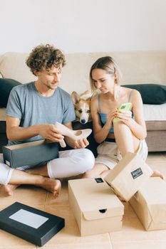Couple sits on the floor surrounded by shoeboxes, with a dog between them, enjoying a cozy moment.