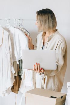 Caucasian woman holding a laptop, selecting clothes from a rack in a fashion studio.