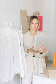 A young woman holding a laptop stands by a clothes rack in a brightly lit room, focused and engaged.