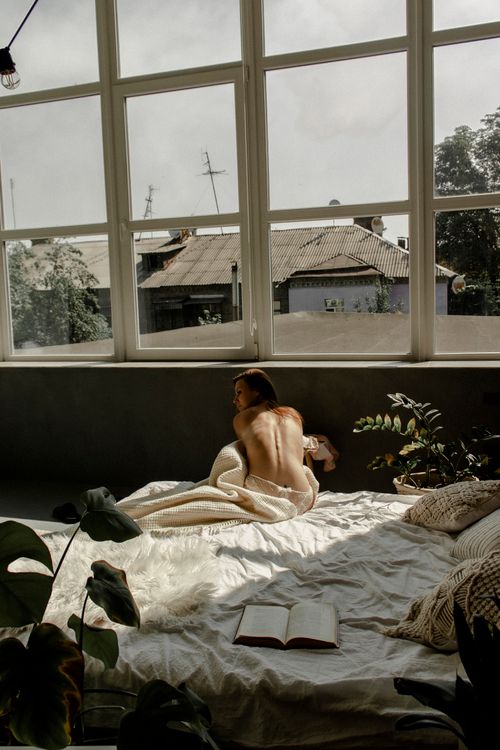 Free Woman sitting on bed in sunlit room, embracing a cozy blanket, with plants and an open book. Stock Photo