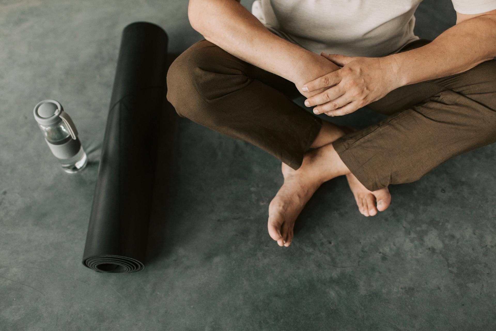 Adult seated on a concrete floor with a rolled yoga mat and water bottle beside him.