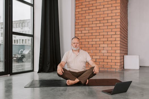 Elderly man practicing meditation sitting cross-legged indoors with a laptop nearby.