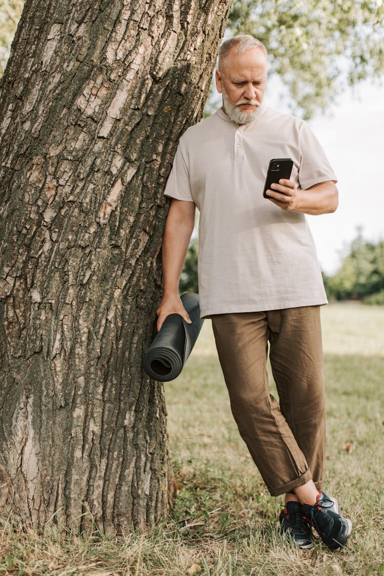 An Elderly Man Leaning On A Tree Trunk While Texting 