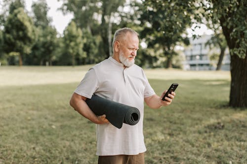 Free Bearded Man Using Cellphone While Holding a Yoga Mat Stock Photo