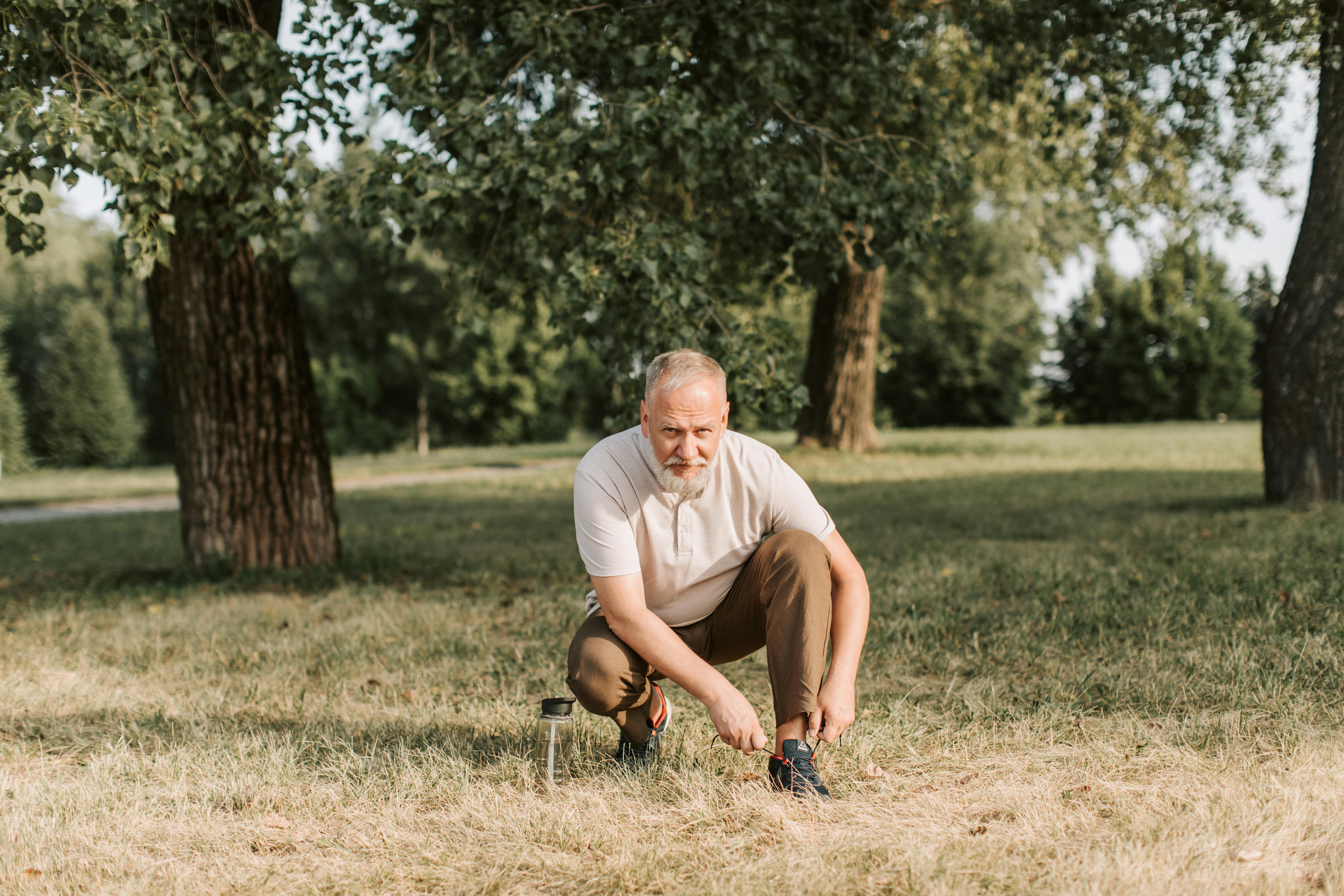 Senior man tying shoelaces in a park, surrounded by grass and trees, enjoying a sunny day