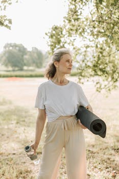 Elderly woman holds yoga mat and water bottle, embracing a healthy lifestyle outdoors.