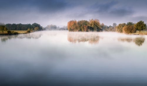 Free Calm autumn lake reflecting colorful trees amidst morning mist under a cloudy sky. Stock Photo