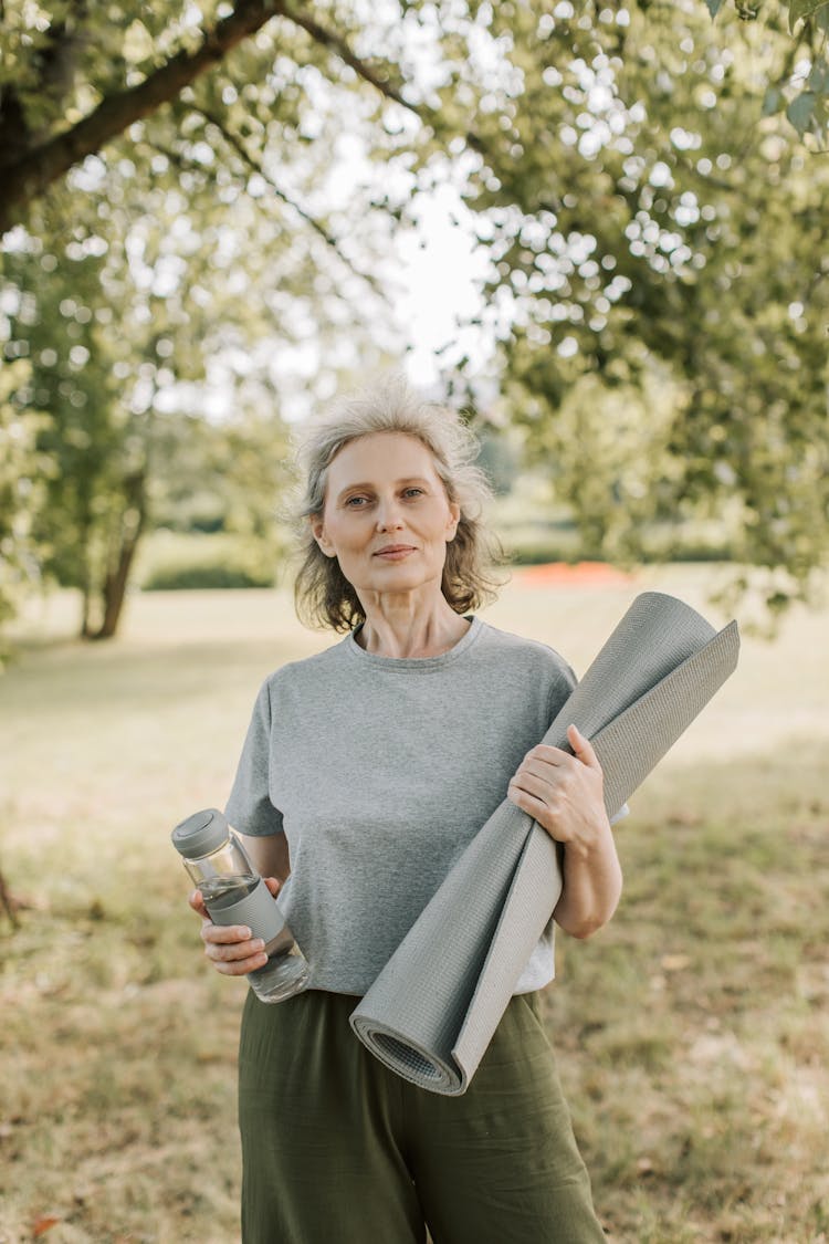 An Elderly Woman Carrying A Yoga Mat While Holding A Bottle Of Water