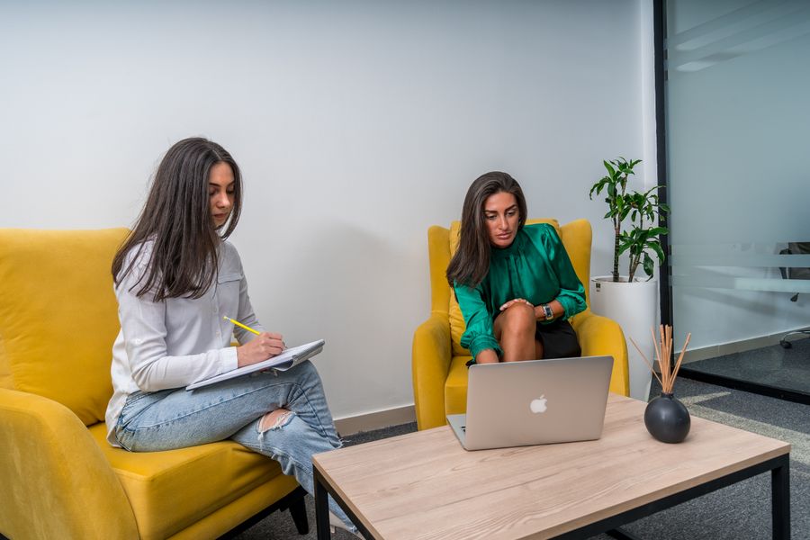 Two women work in a modern office setting, seated on yellow chairs, focusing on a laptop and notes.