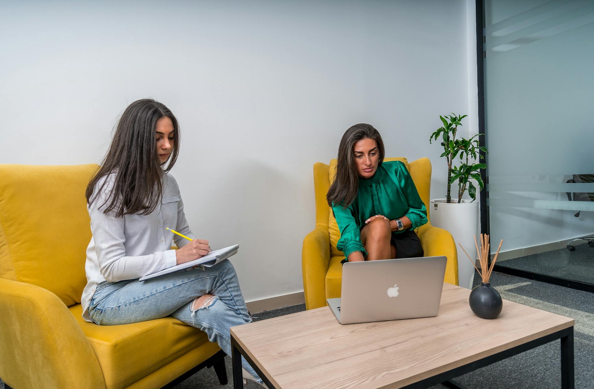 Two women work in a modern office setting, seated on yellow chairs, focusing on a laptop and notes.