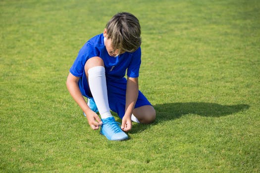 A child in a blue soccer uniform tying shoelaces on a grassy field in daylight.