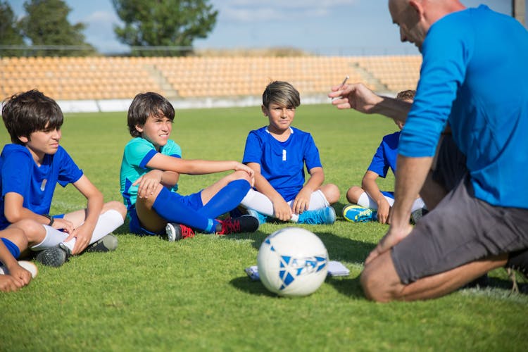 A Man In Blue Shirt Coaching A Group Of Boys Sitting On A Soccer Field