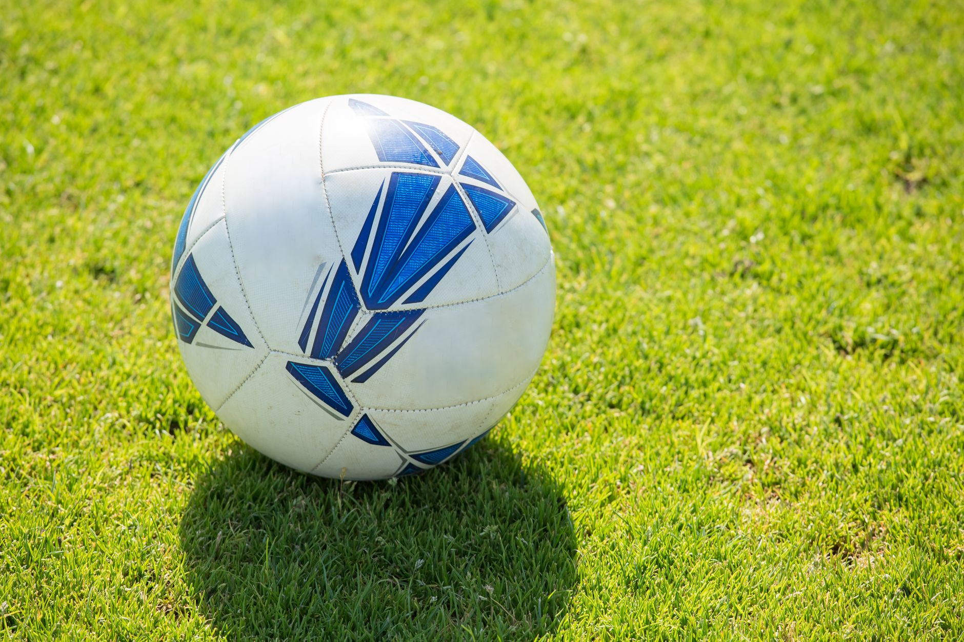 Bola sepak putih dengan pola biru yang terletak di lapangan rumput hijau di bawah sinar matahari.