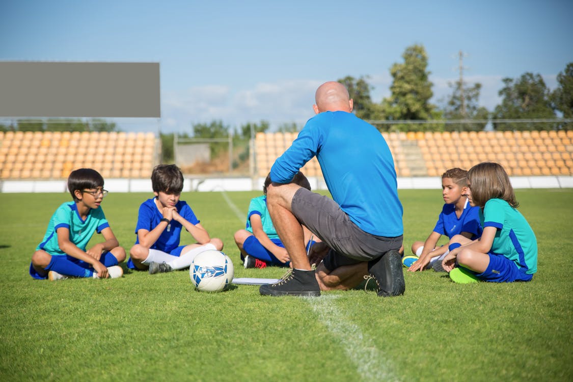 Ein Mann Im Blauen Hemd Trainiert Eine Gruppe Von Kindern, Die Auf Einem Fußballfeld Sitzen