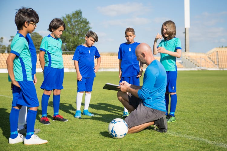 A Man In Blue Shirt Coaching A Group Of Kids Standing On A Soccer Field