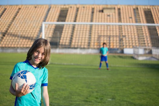 Two kids enjoying a game of soccer at an outdoor field with a stadium backdrop.