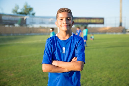Smiling boy in blue jersey standing confidently on a soccer field during sunset.