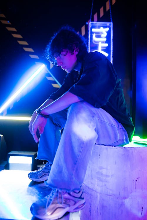 Young Man in a Trendy Outfit Sitting in a Room with Neon Lights 
