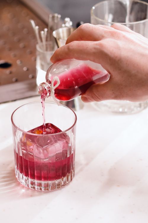 Free Close-up of hand pouring vibrant red cocktail into glass with large ice cube on a bar counter. Stock Photo